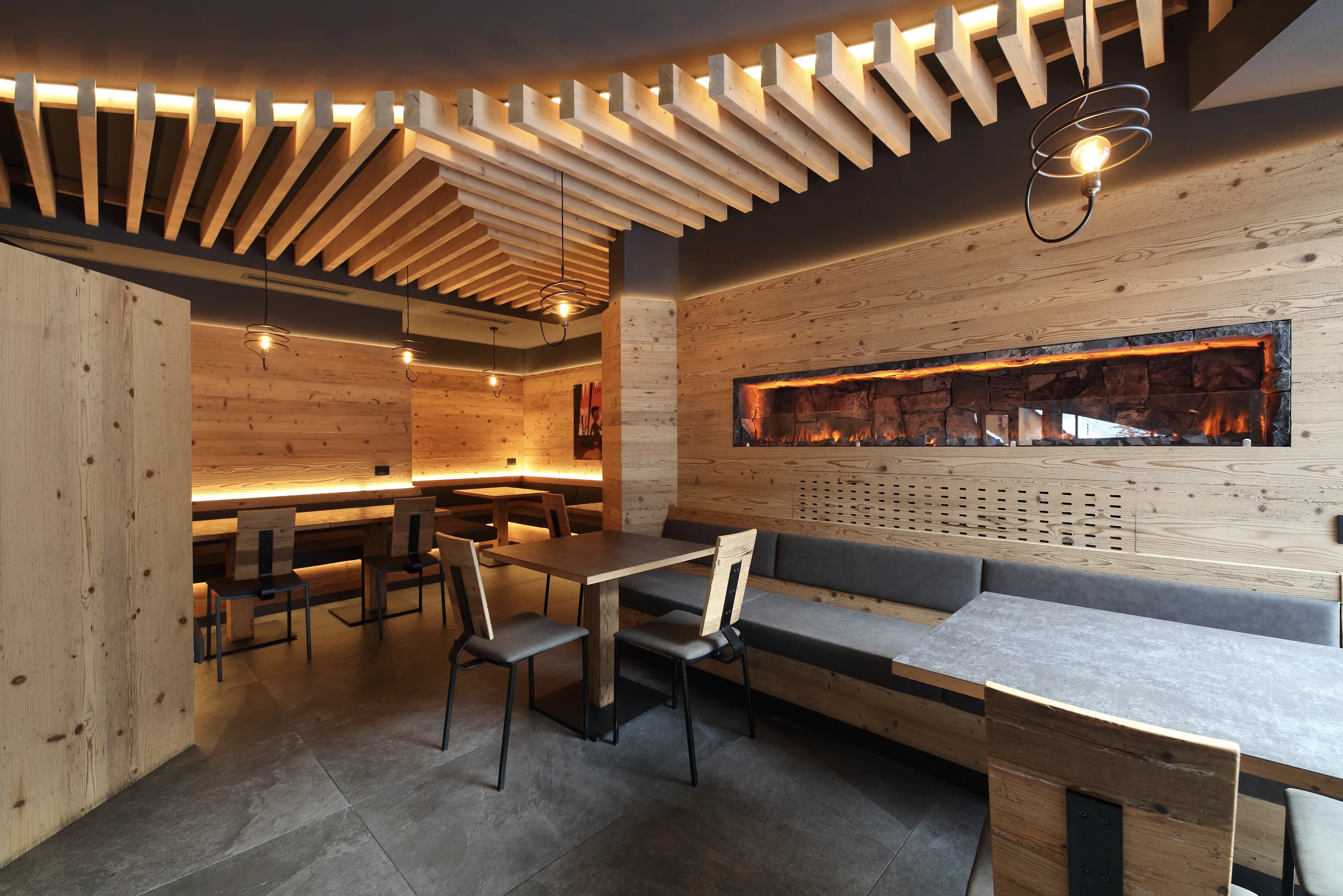 Interno di un ristorante moderno in legno con soffitto a listelli, tavoli, panche imbottite e camino lineare integrato.
