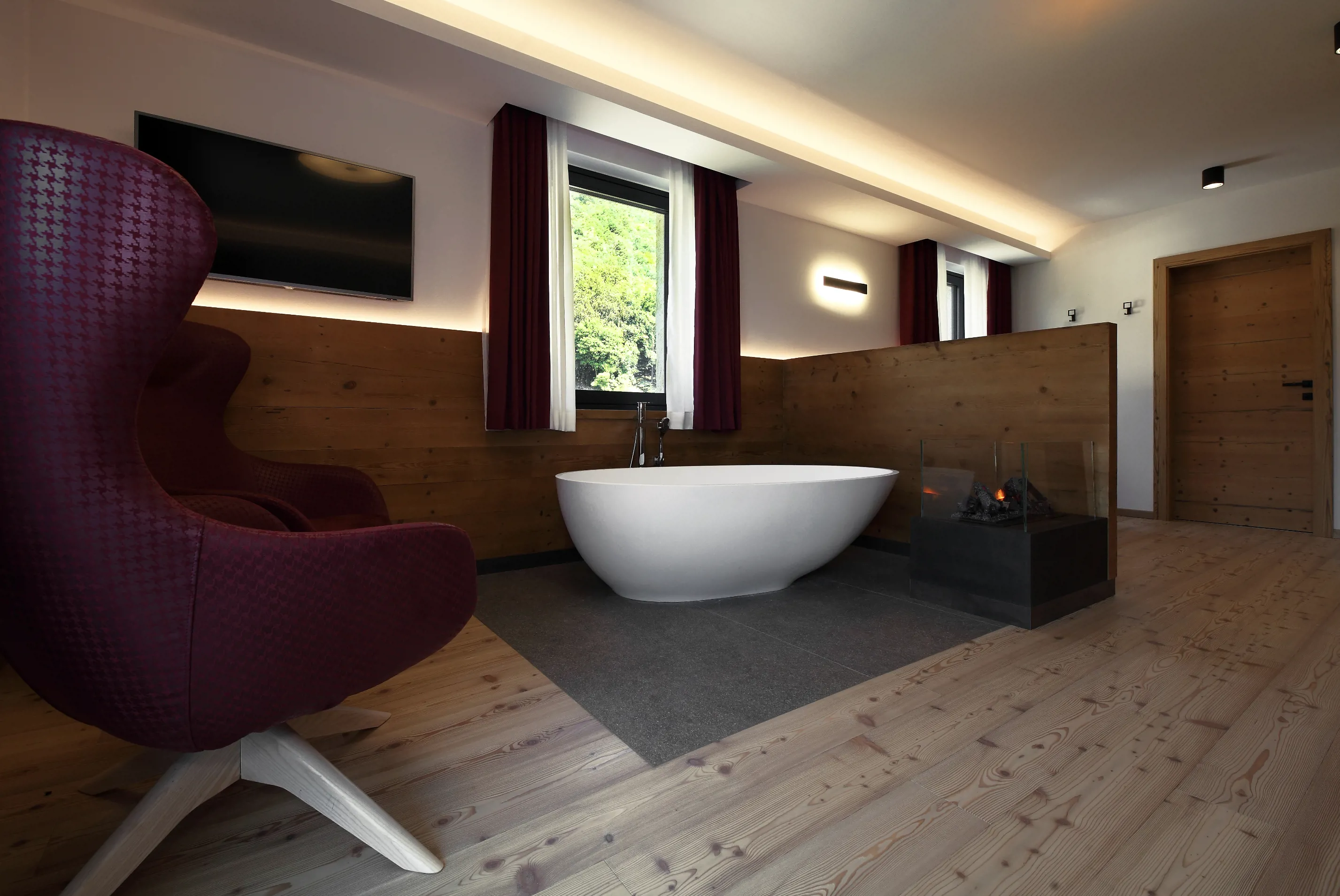 Moderna camera d'albergo con vasca autoportante bianca, pannelli in legno, poltrona bordeaux e camino in vetro.